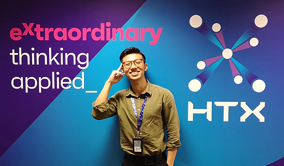 A smiling man with glasses and a lanyard poses in front of a colorful HTX branded wall reading "extraordinary thinking applied."